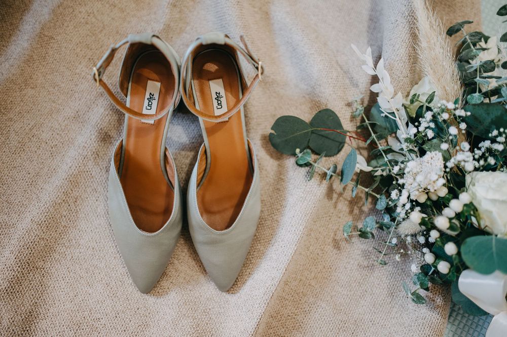 Gray bridal shoes displayed next to white and green floral arrangement on beige fabric.