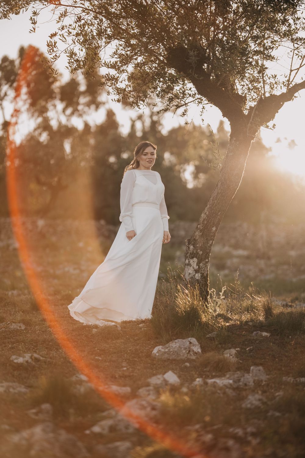 Person in flowing white dress stands beneath tree during golden sunset.