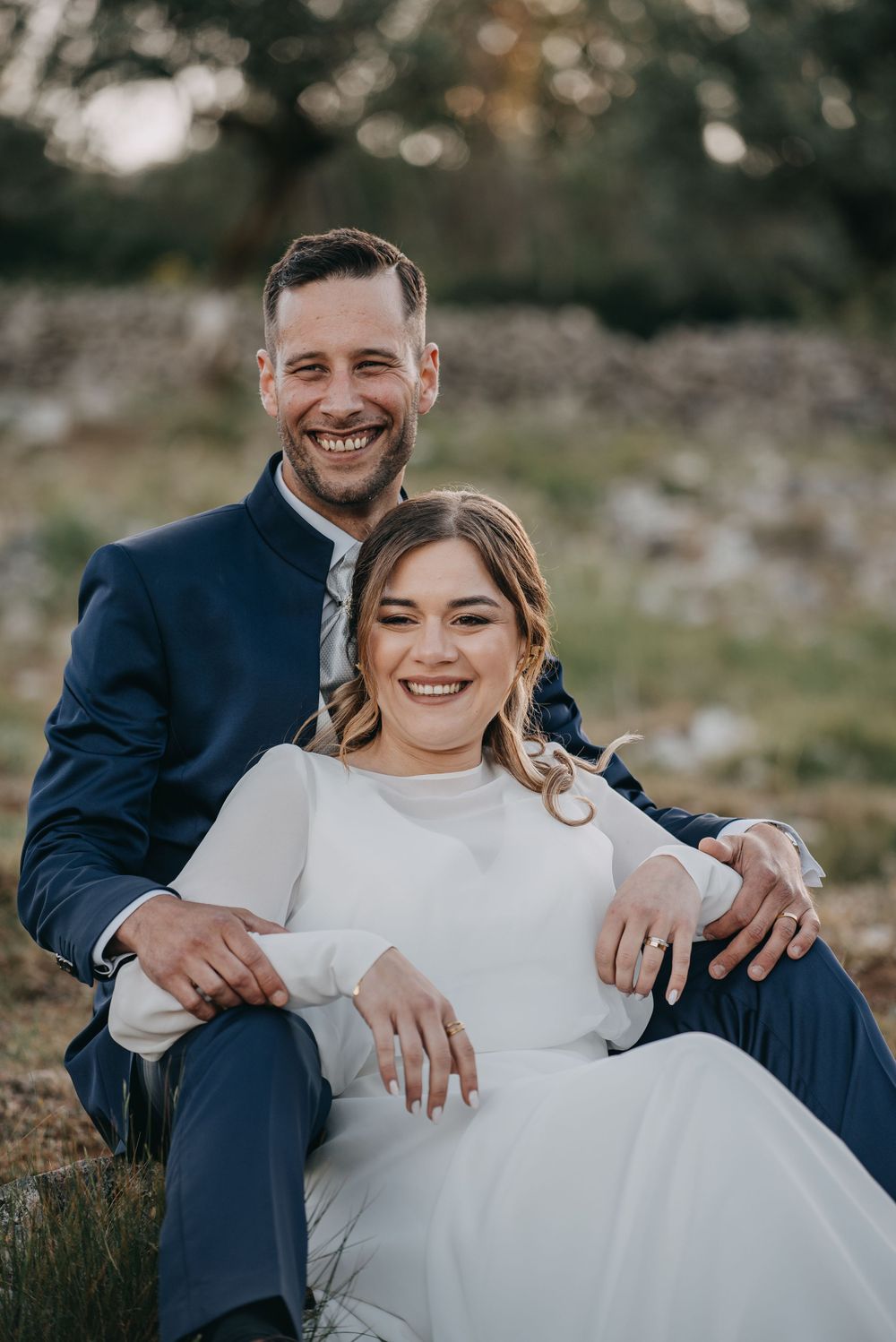 A joyful wedding portrait of a couple in formal attire sitting on natural terrain.