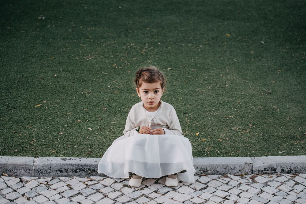 A small child in white dress sitting on stone pavement against green grass background.