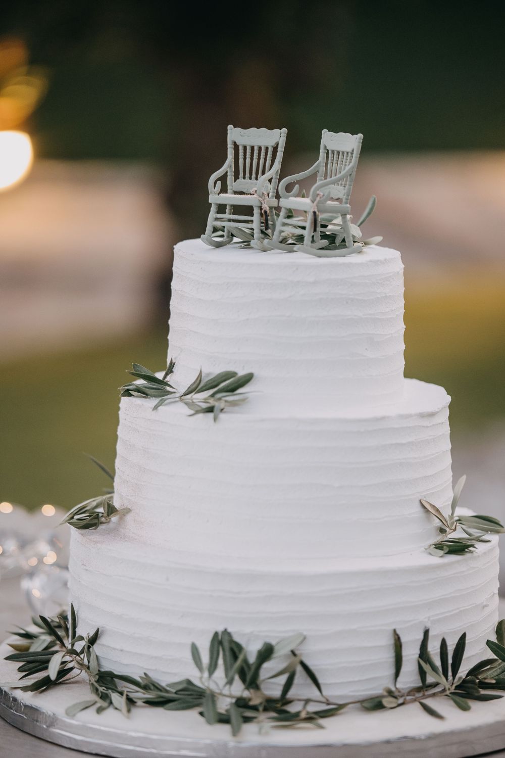 White tiered wedding cake decorated with olive leaves and miniature rocking chair toppers.