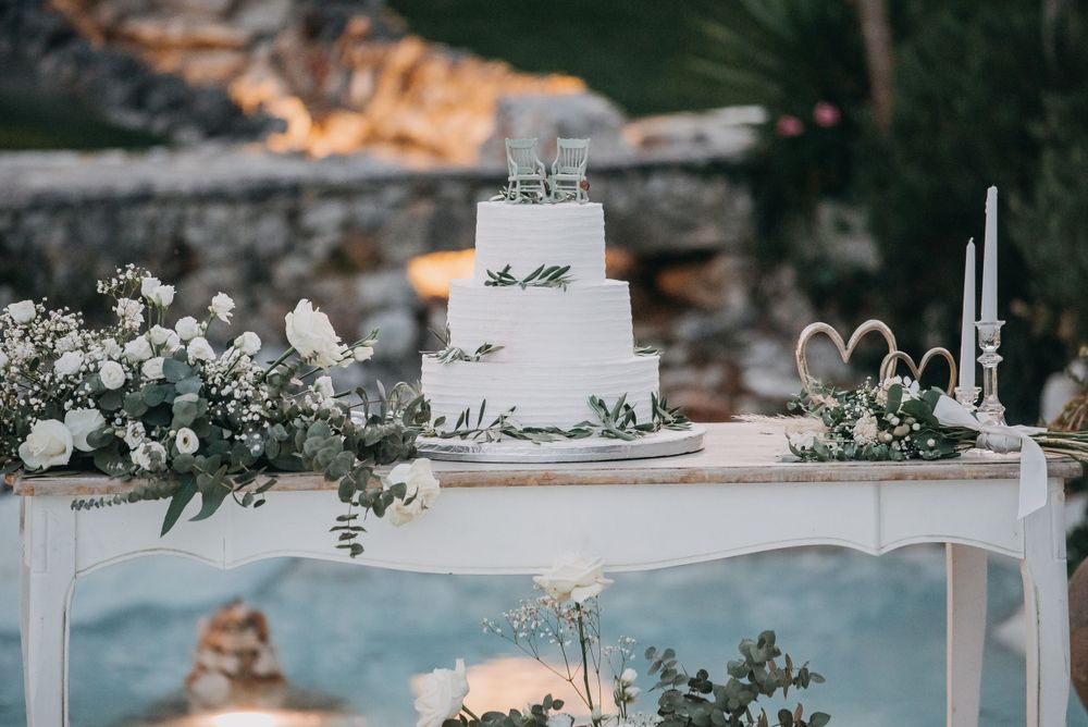 Elegant white wedding cake displayed on a white table with greenery and floral decorations.