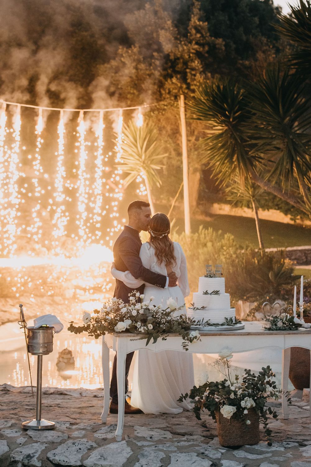 Wedding cake cutting moment against a backdrop of cascading sparkler lights at sunset.