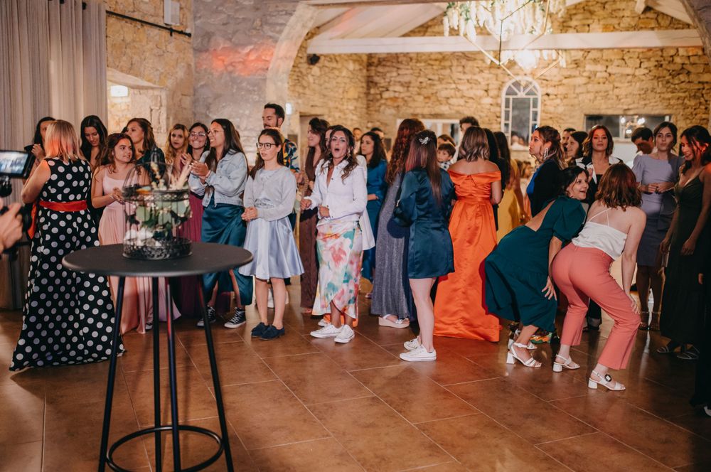Wedding guests in colorful attire dance and celebrate in a rustic stone-walled venue.
