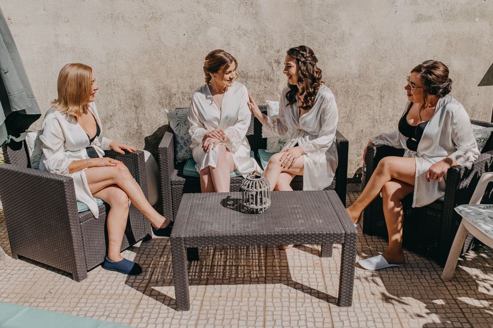 Four women in matching robes sitting outdoors on patio furniture having conversation.