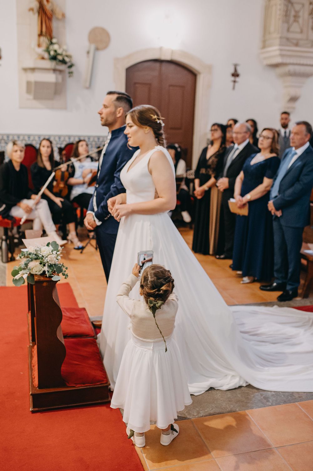A wedding ceremony in a church with people standing on a red carpet while a small child tends to the bride's train.