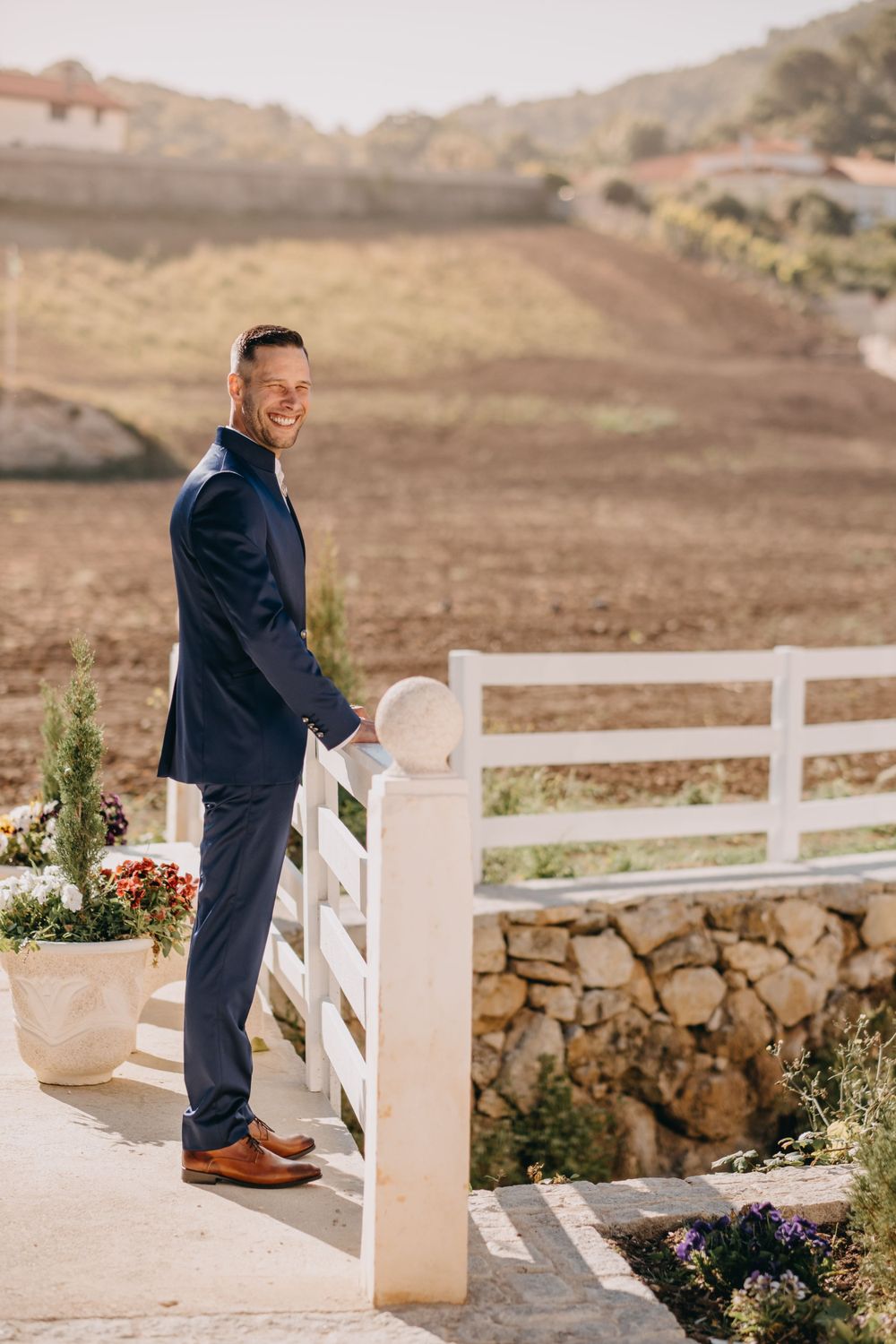 Person in navy suit standing by white fence with potted flowers and hillside landscape.