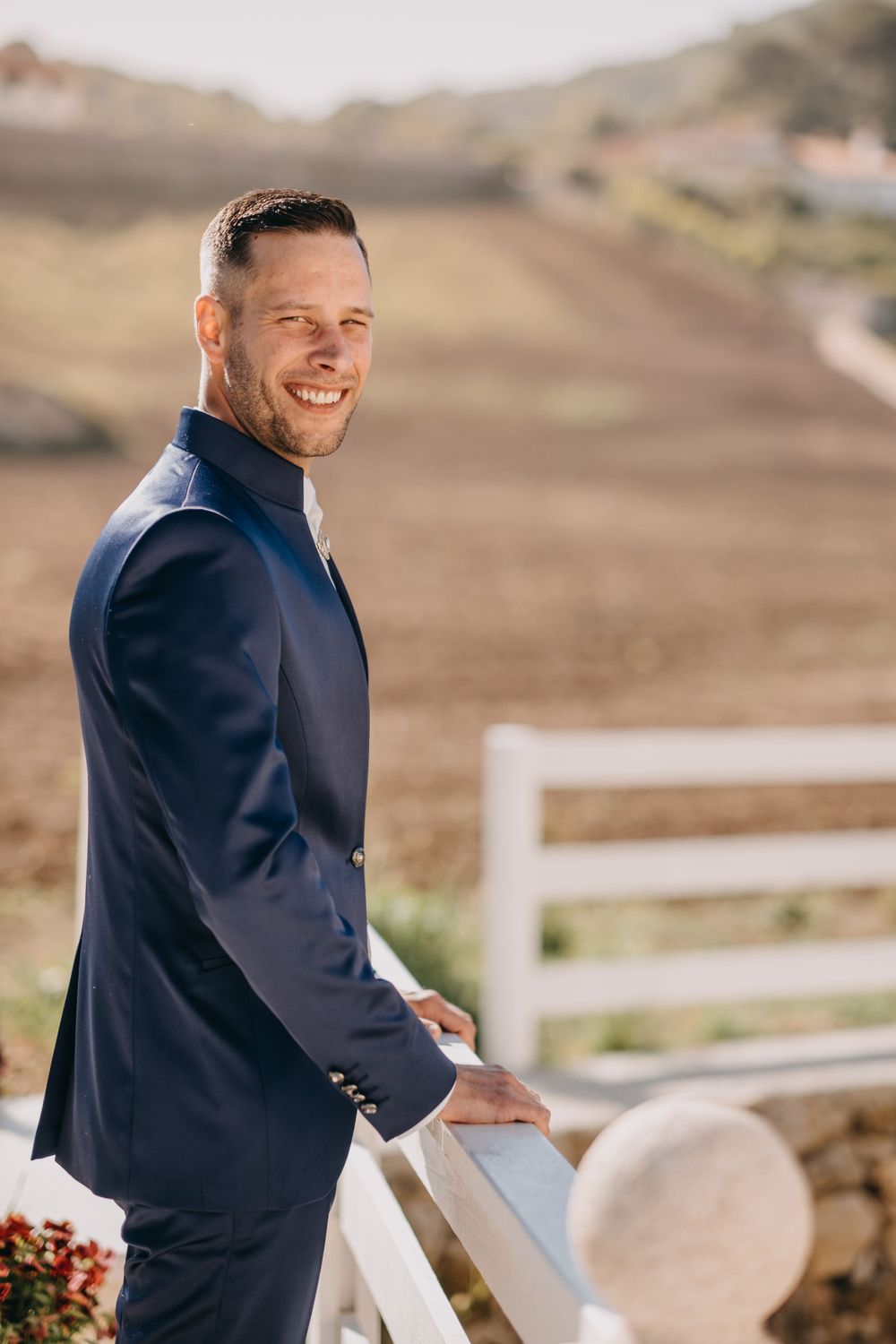 Person in formal attire smiling by white fence with scenic countryside background.