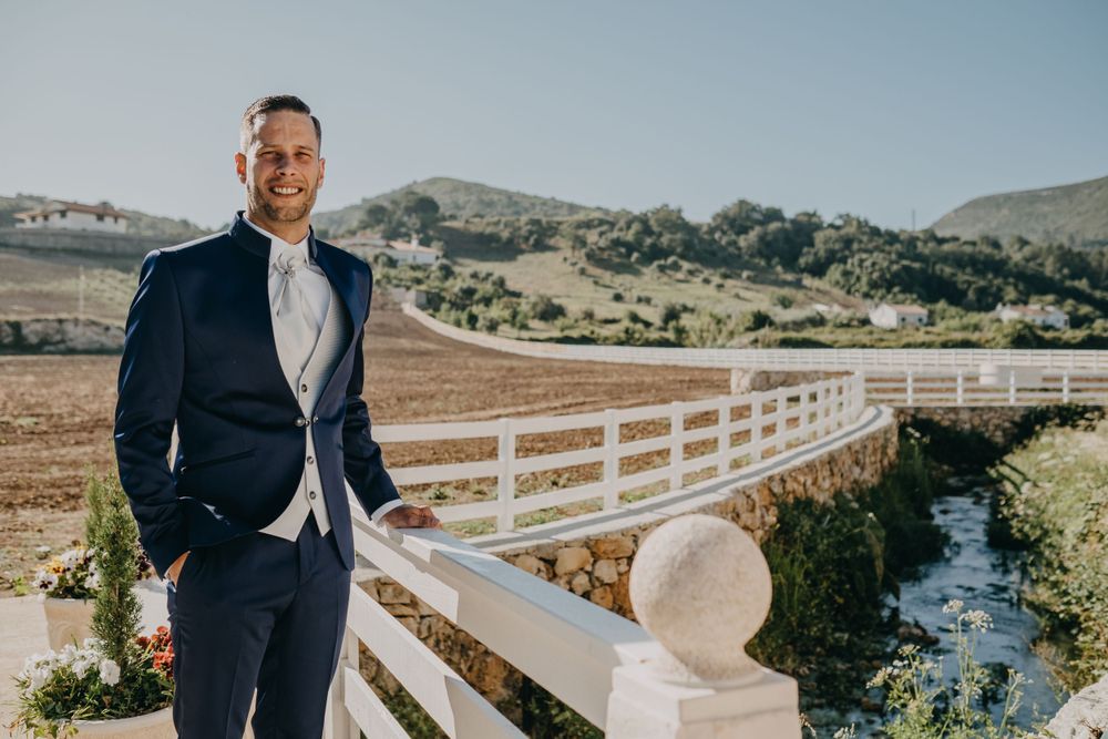 Person in dark suit posing against white fencing with rolling hills in background.