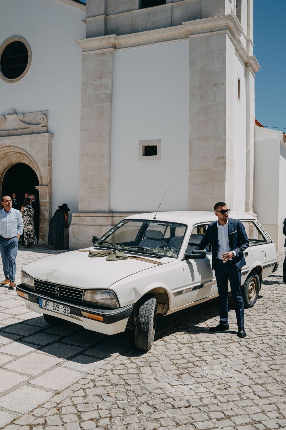A vintage white car parked outside a white church building with cobblestone pavement.
