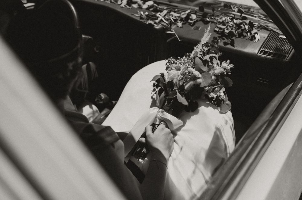Black and white view through car window showing bridal bouquet and wedding dress details.