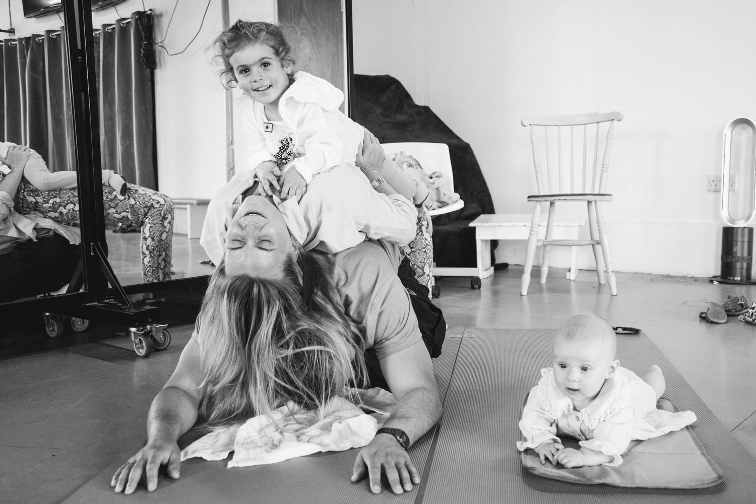 Black and white photo of participants doing stretching exercises in a studio with baby nearby.