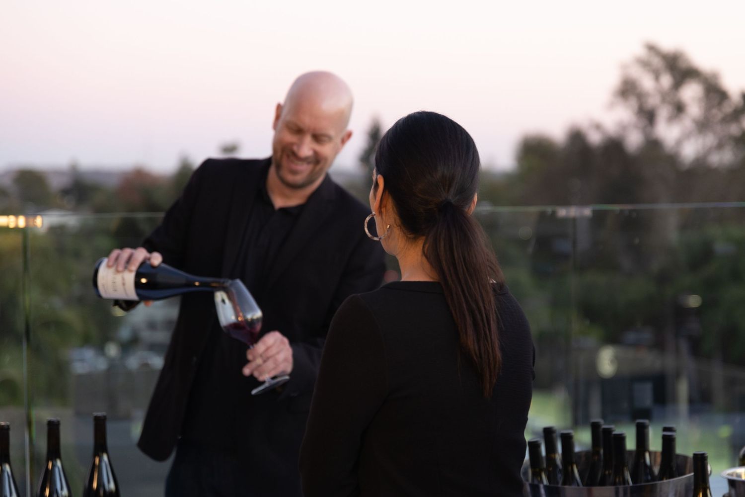 A smiling bartender pours a glass of wine for a woman at a corporate tasting event while the sun sets in the distance.