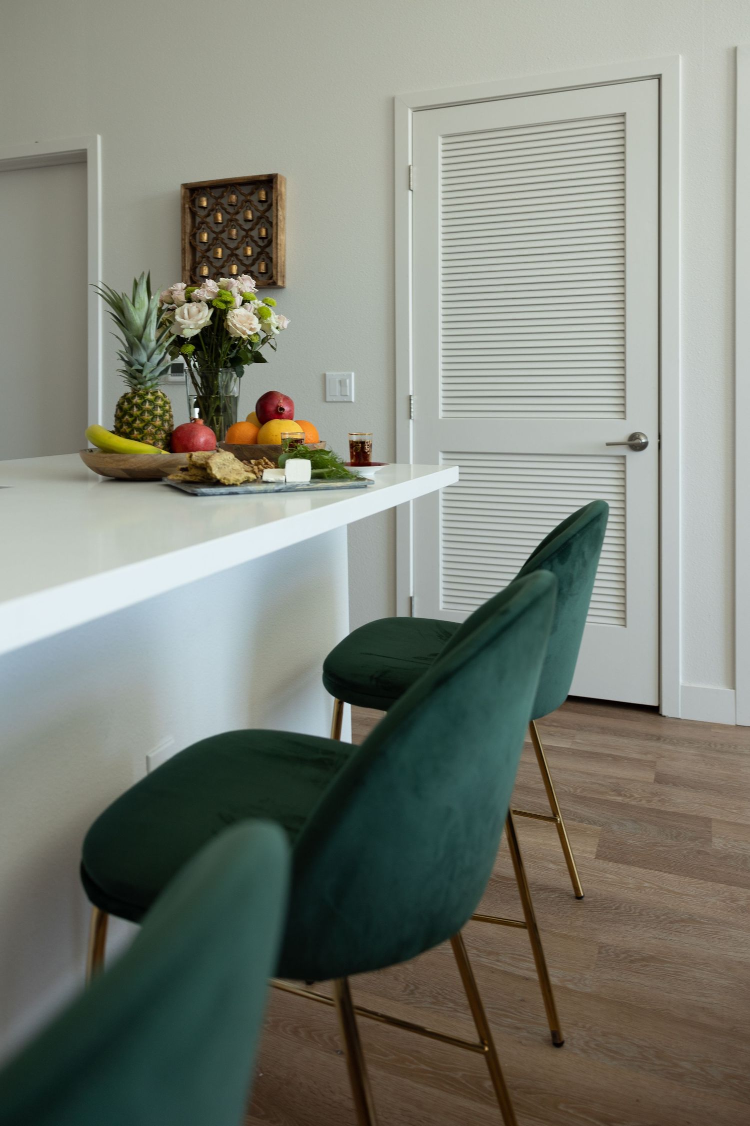 Home interior featuring jewel-tone green velvet chairs along a white kitchen bar counter decorated with flowers and fruit.