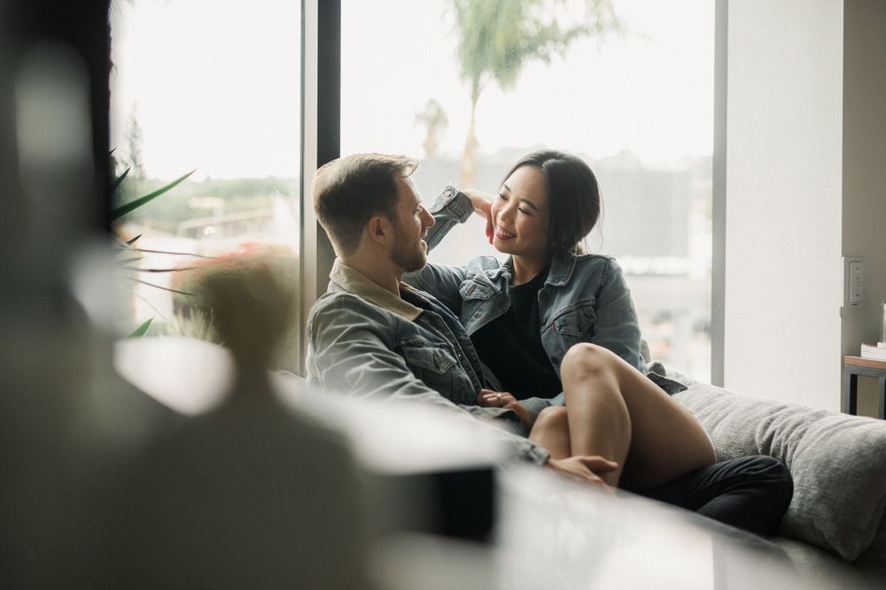 Couple sharing intimate moment on grey couch by window with plants in background.