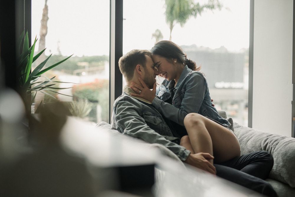 Couple cuddling on couch by window with indoor plants visible in modern interior setting.
