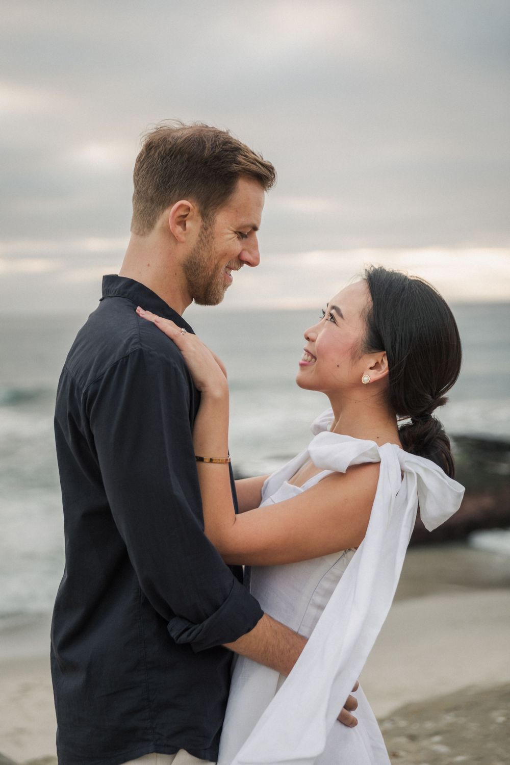 A romantic couple embraces on a beach with rocky coastline and cloudy skies in the background.
