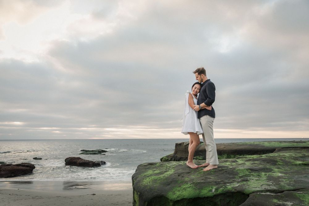 A couple stands on a moss-covered rock overlooking the ocean during sunset.