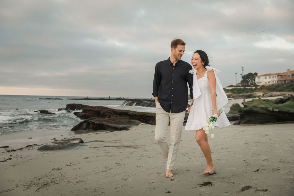 A couple strolls hand in hand on a sandy beach with rocks and ocean waves in the background.
