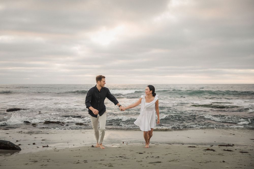 A couple holds hands while walking barefoot along a moody beach shoreline.