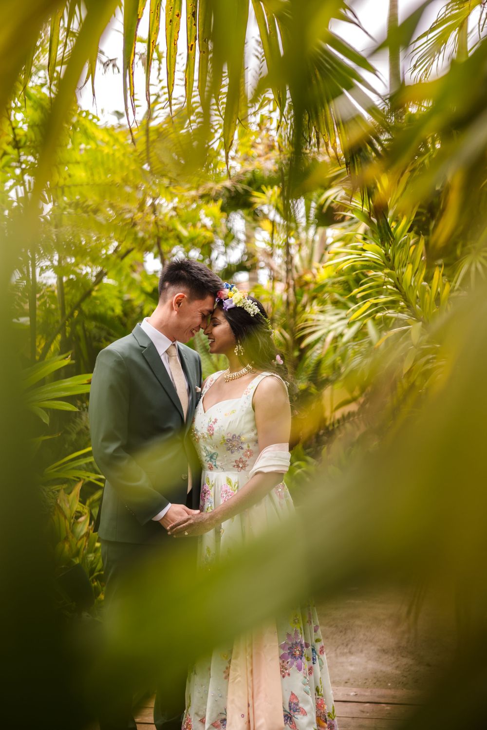 A romantic wedding photo through palm fronds showing a couple in formal attire embracing.