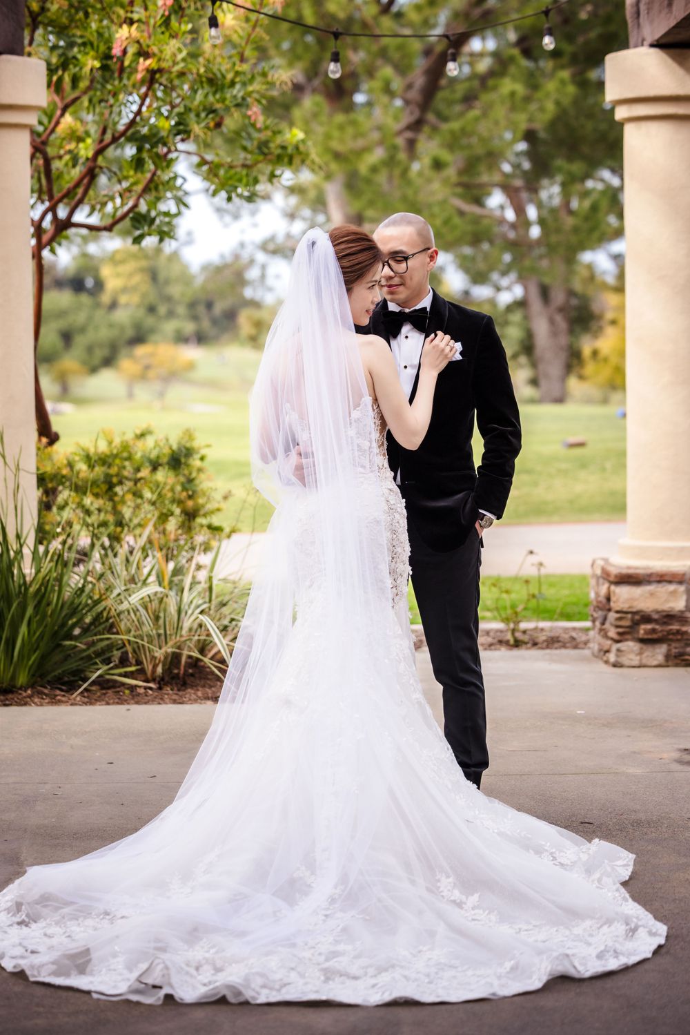 An elegant wedding couple poses between stone pillars as her lace train spreads across the ground.