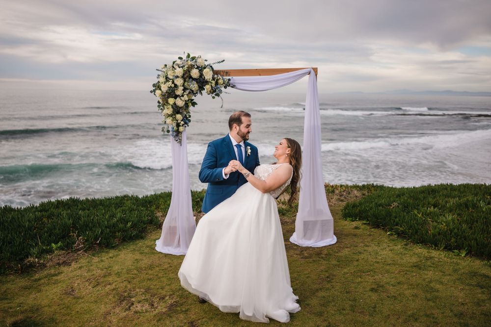 Wedding ceremony setup on coastal cliff with white arch and floral arrangements overlooking ocean.