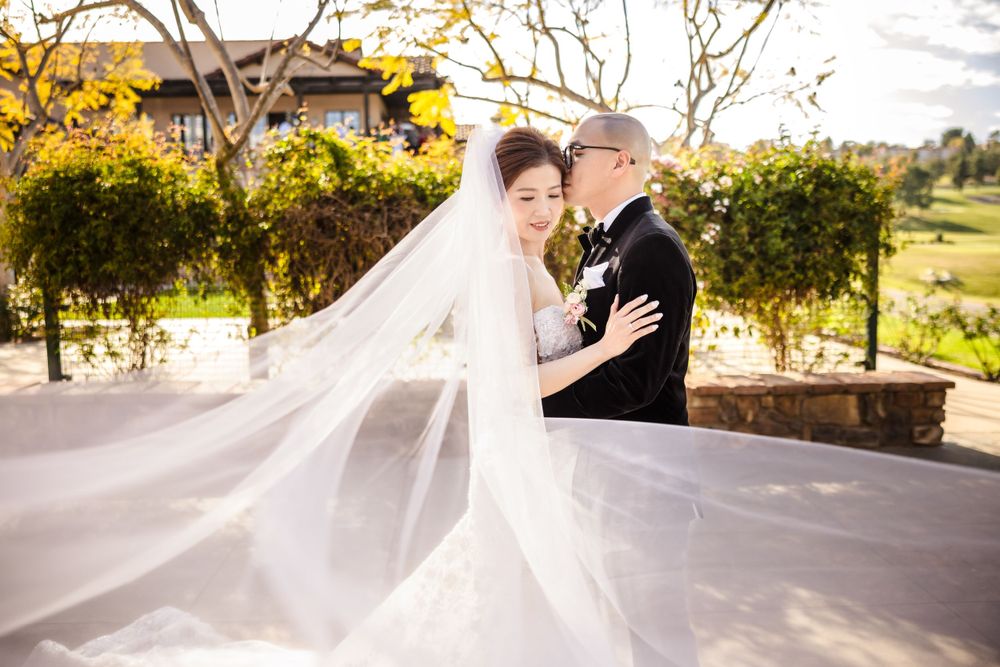 A flowing veil catches the sunlight as a couple shares an intimate moment in an outdoor garden setting.