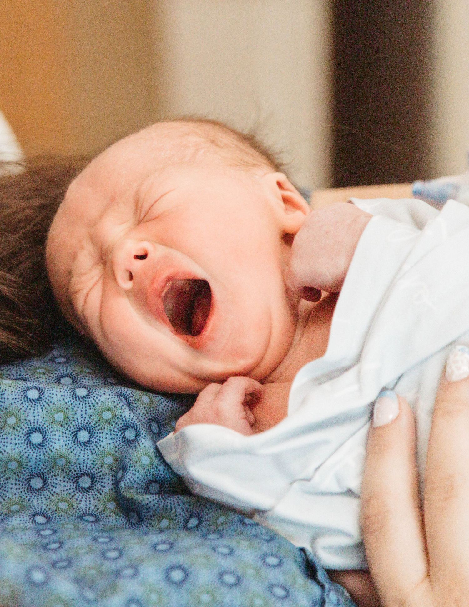 A sleepy baby yawns while lying on a blue polka dot pillow, wearing a light colored outfit.