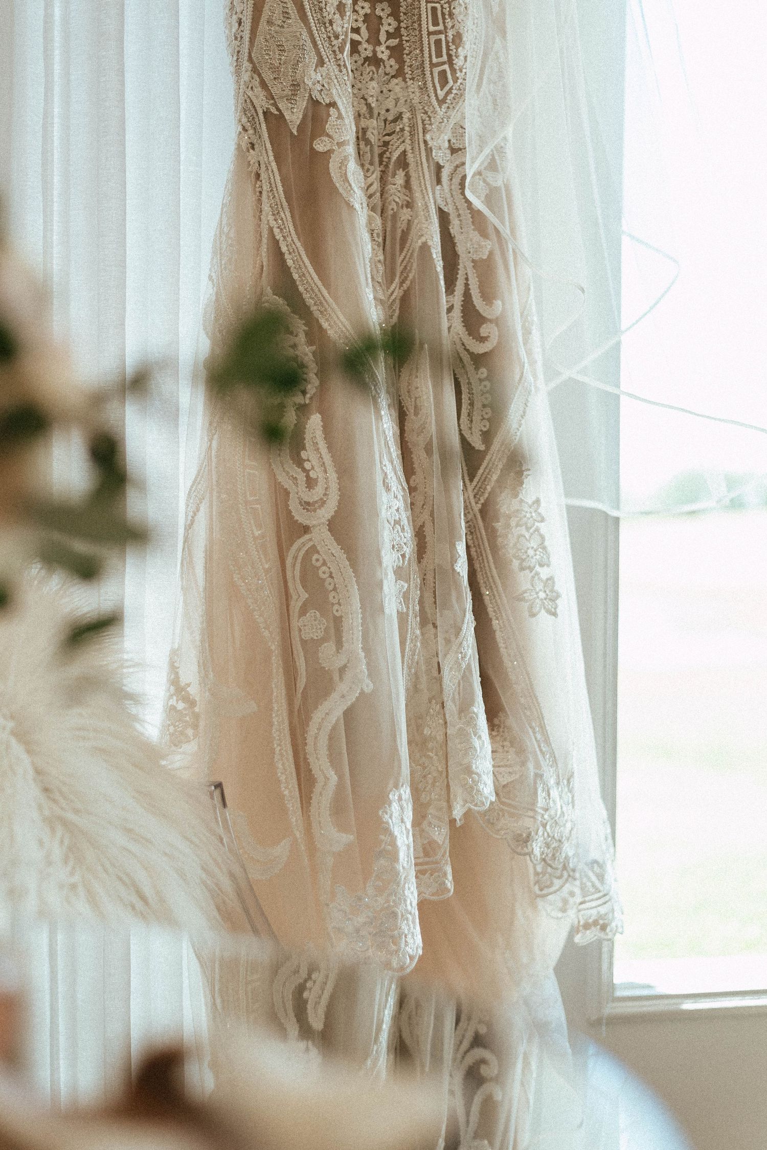 Close-up detail of a delicate lace wedding dress with intricate beading hanging by a window.