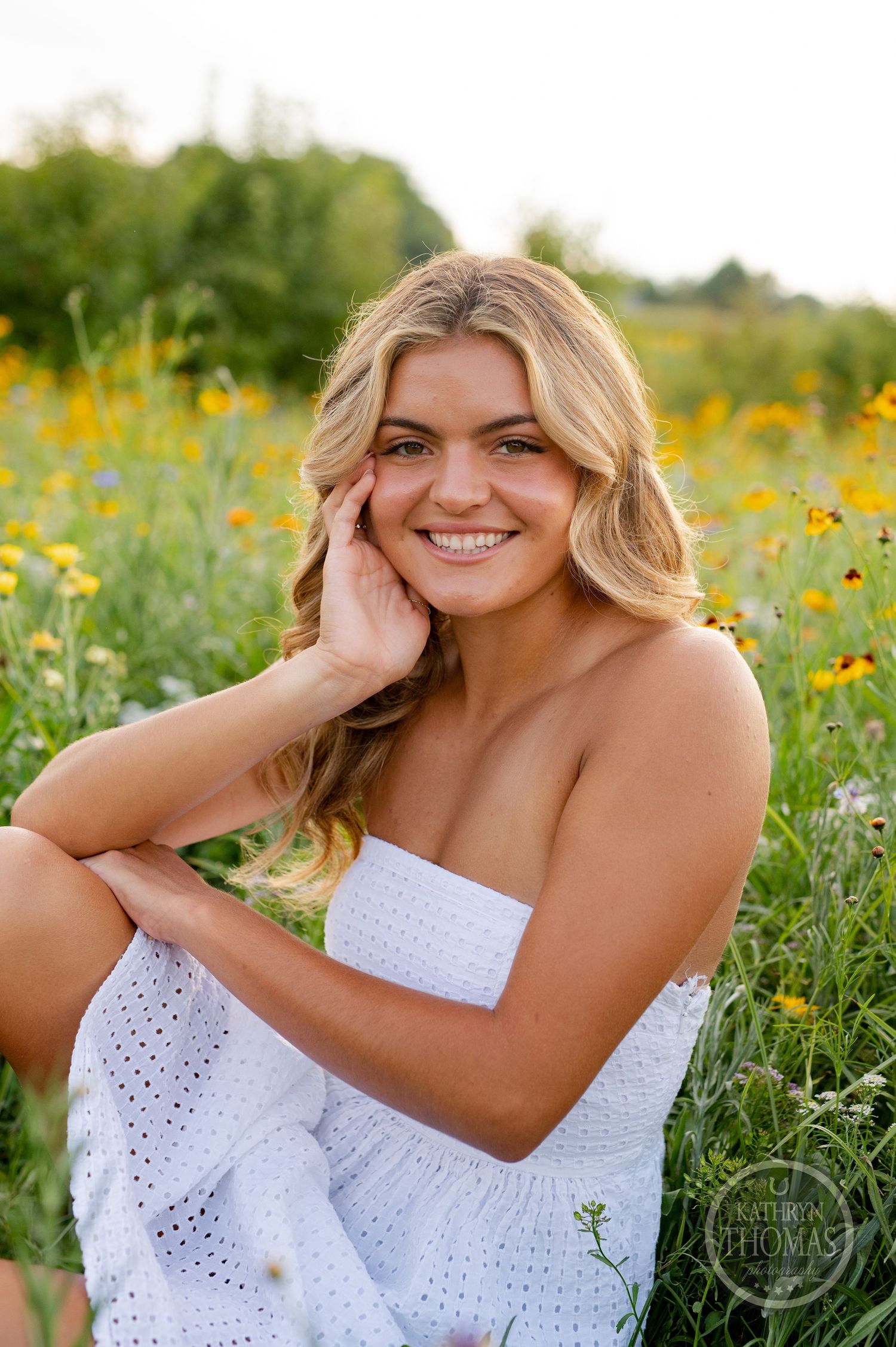 A woman in a white strapless dress smiles warmly while sitting in a blooming wildflower meadow.