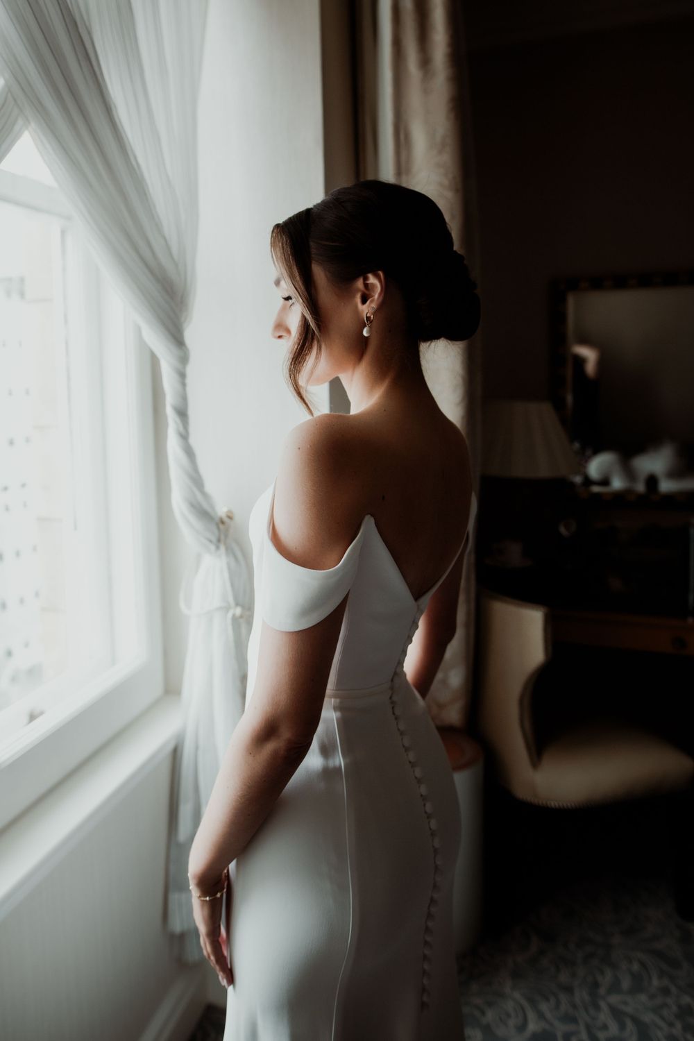 Elegant silhouette of bride in a white gown looking out a window in soft natural light London wedding photographer. The landmark