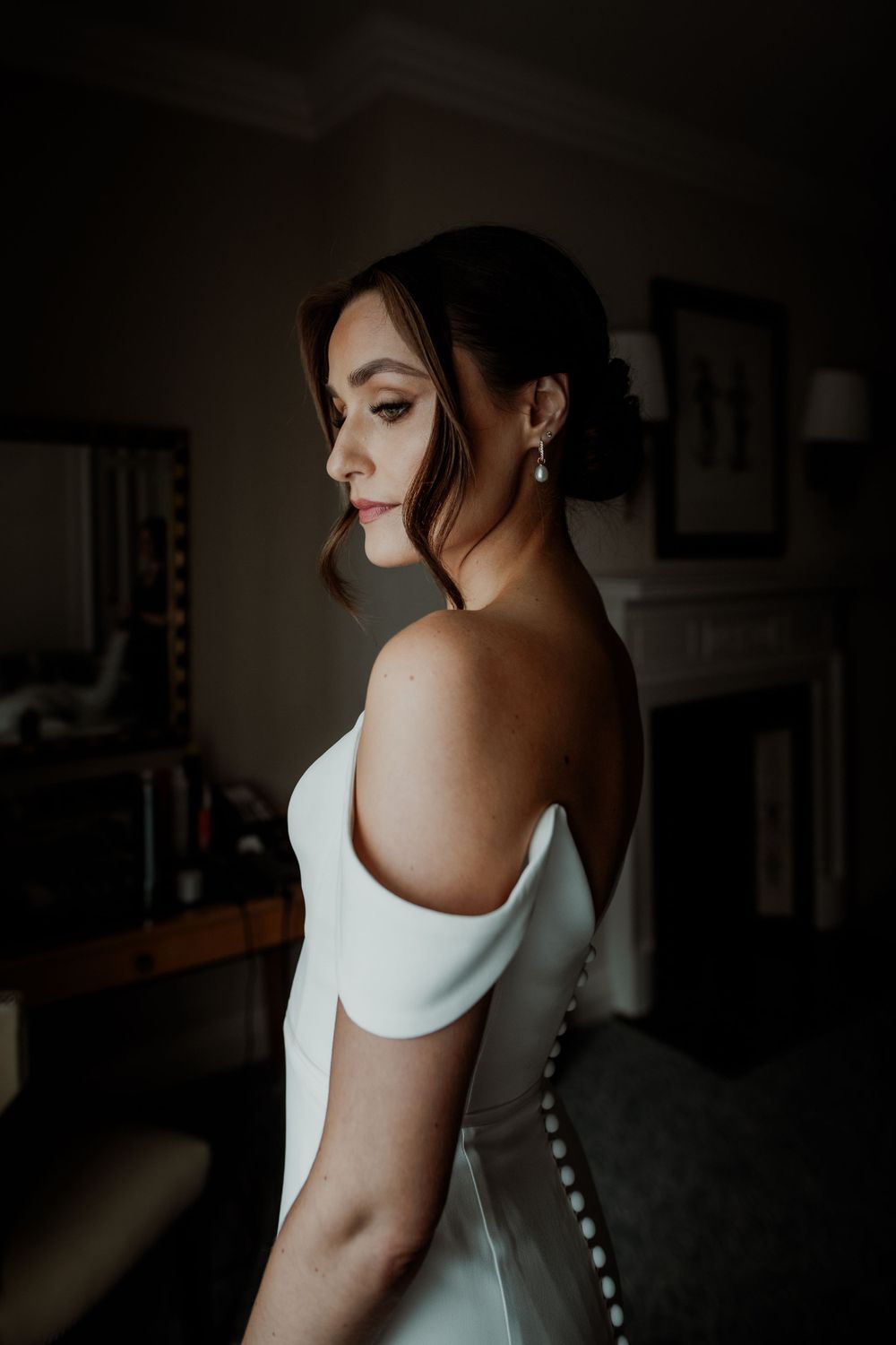 Close-up profile view of someone wearing an elegant white off-shoulder wedding dress in a dimly lit room. London wedding photographer