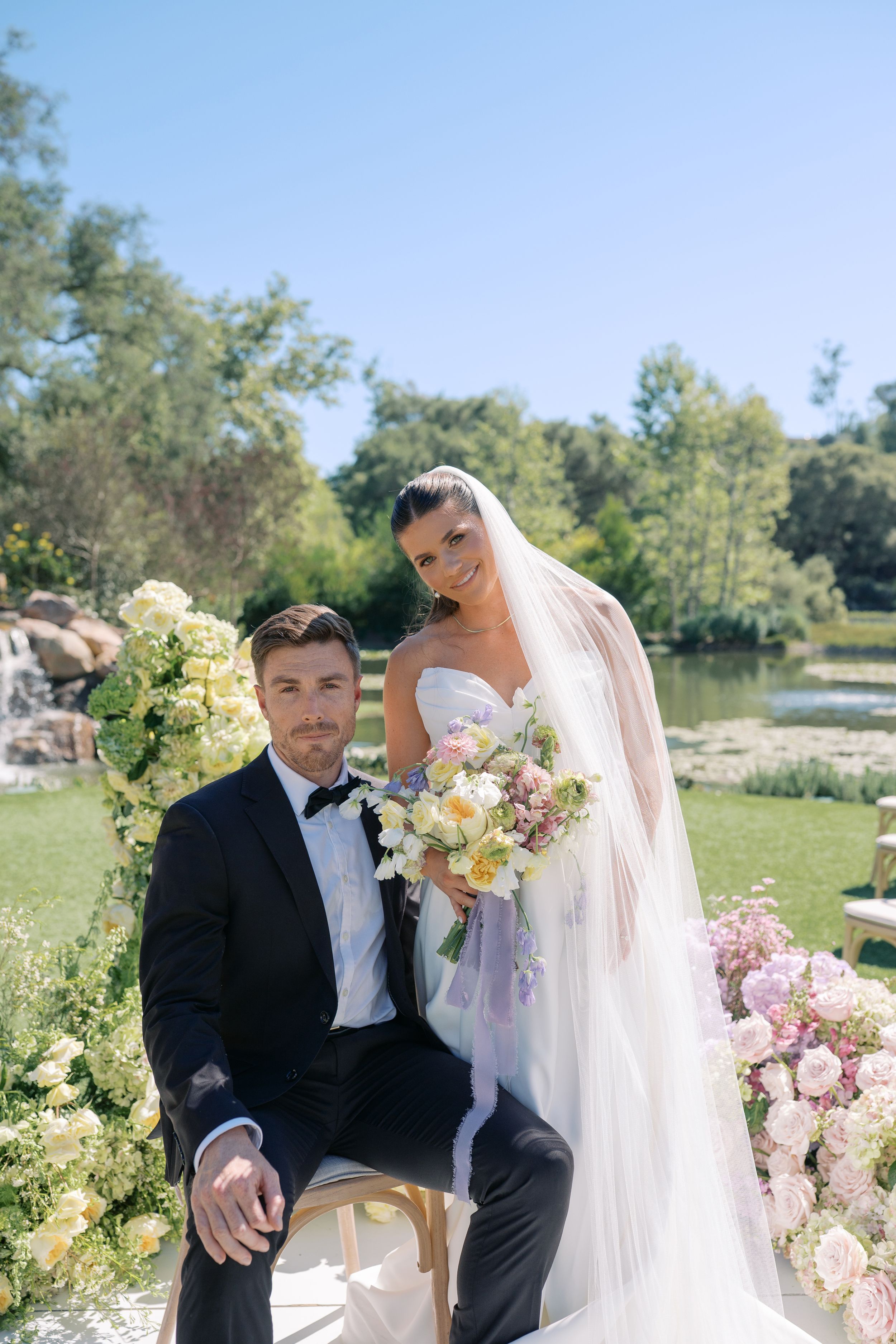 A romantic outdoor wedding scene with a couple sitting together surrounded by pink and yellow flowers in a garden setting.