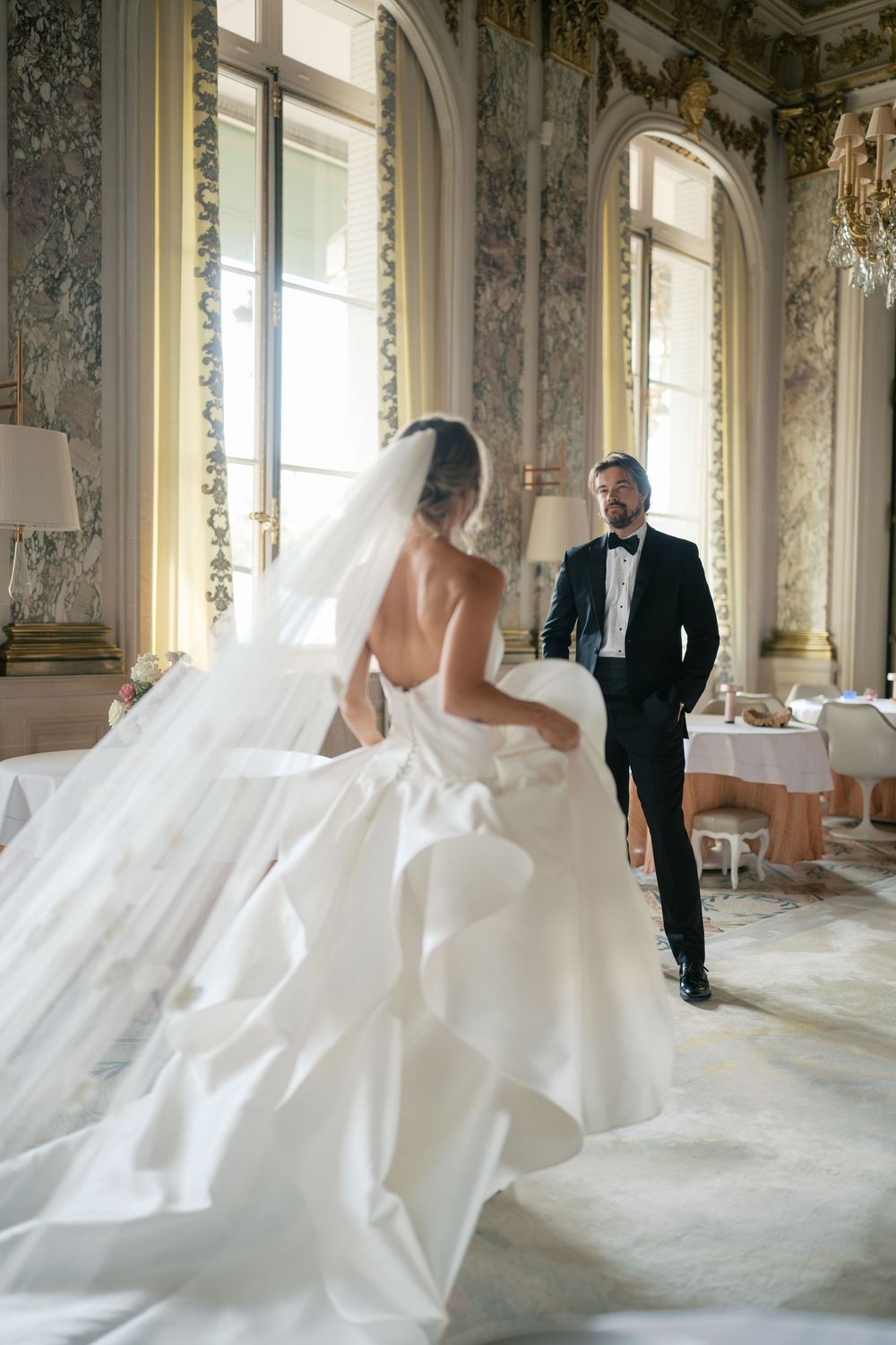 A bride in a flowing white ballgown and long veil faces a groom in a tuxedo inside an ornate classical ballroom.