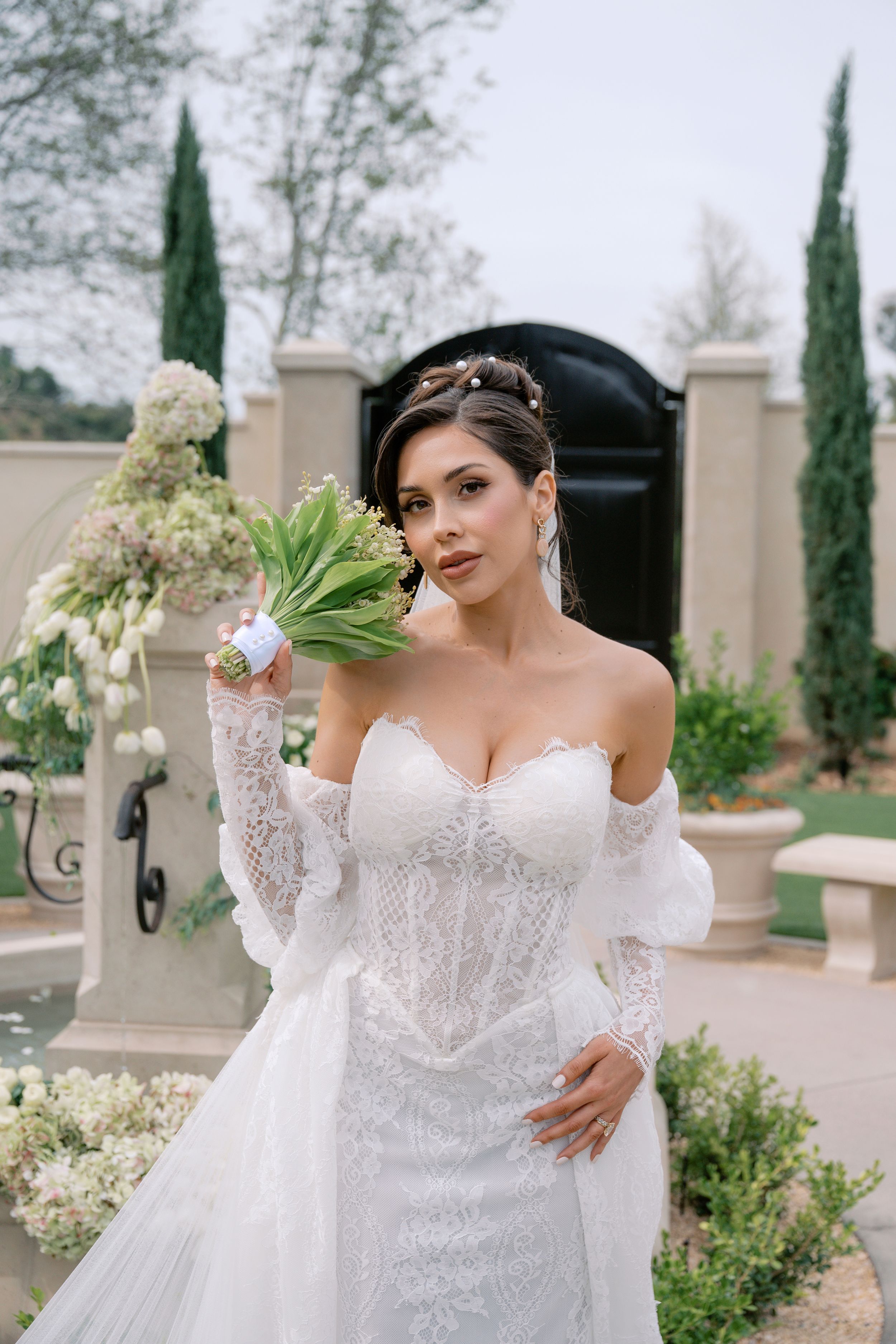 Bride in white lace off-shoulder gown holds green and white bouquet outdoors near elegant stone gate.