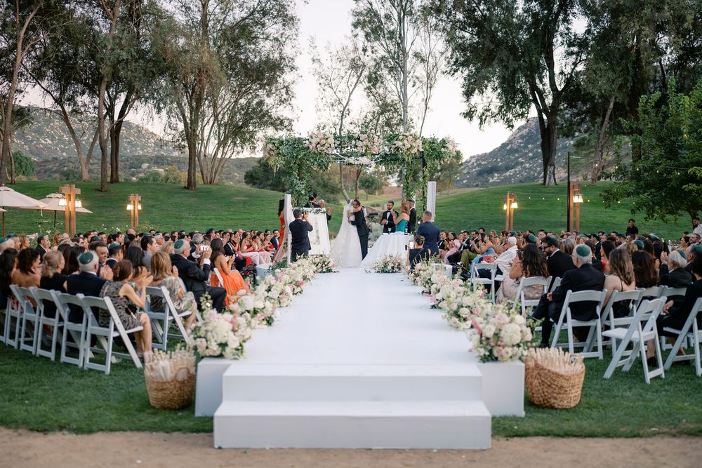 Elegant outdoor wedding ceremony on white aisle runner surrounded by floral arrangements and guests seated in white chairs.