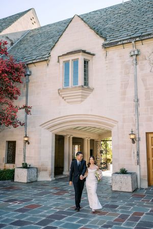 Bride and groom walk hand-in-hand outside an elegant stone mansion with arched entry and blooming bougainvillea.