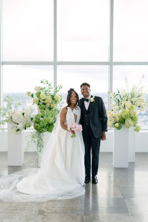 A wedding couple walks down a bright white venue hallway decorated with floral arrangements on pedestals and large windows.
