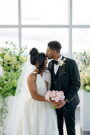 Elegant couple in wedding attire share an intimate moment by bright windows surrounded by green and white floral decor.