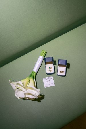 White calla lily bouquet beside two blue jewelry boxes with gold rings on a green background.