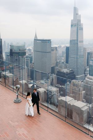 Bride and groom stand on a rooftop observation deck overlooking the iconic New York City skyline on a cloudy day.