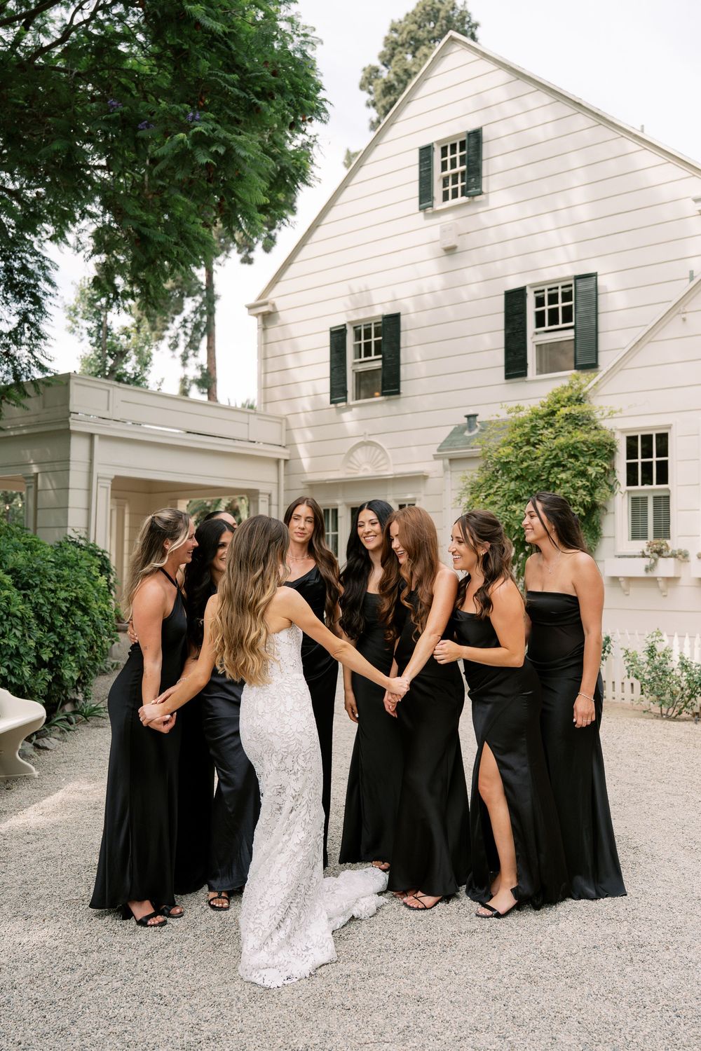 Bride in white lace gown surrounded by bridesmaids in black dresses holding hands outside elegant white estate.