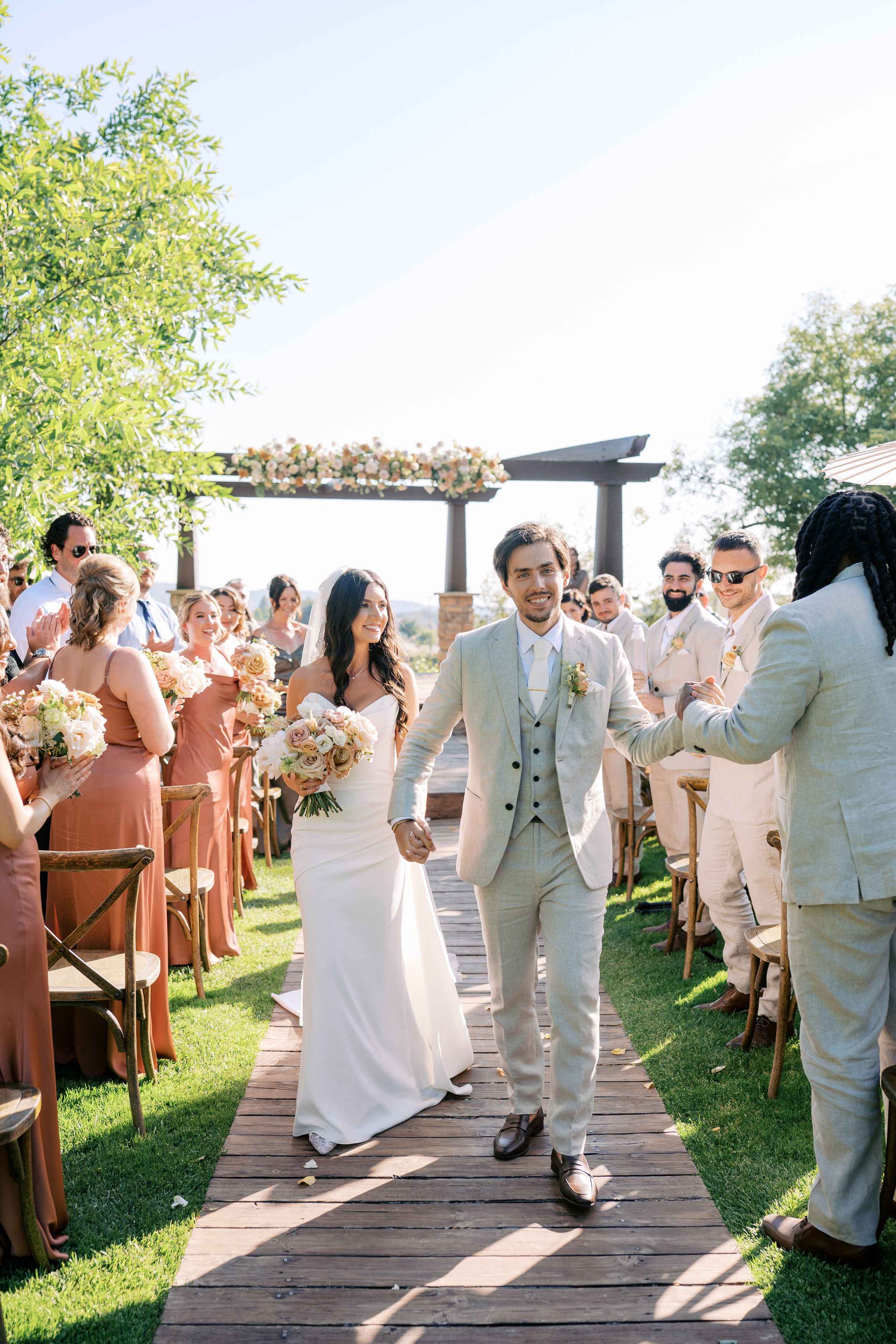 Bride and groom walk down outdoor wedding aisle, guests in terracotta dresses, floral arch backdrop, sunny ceremony.