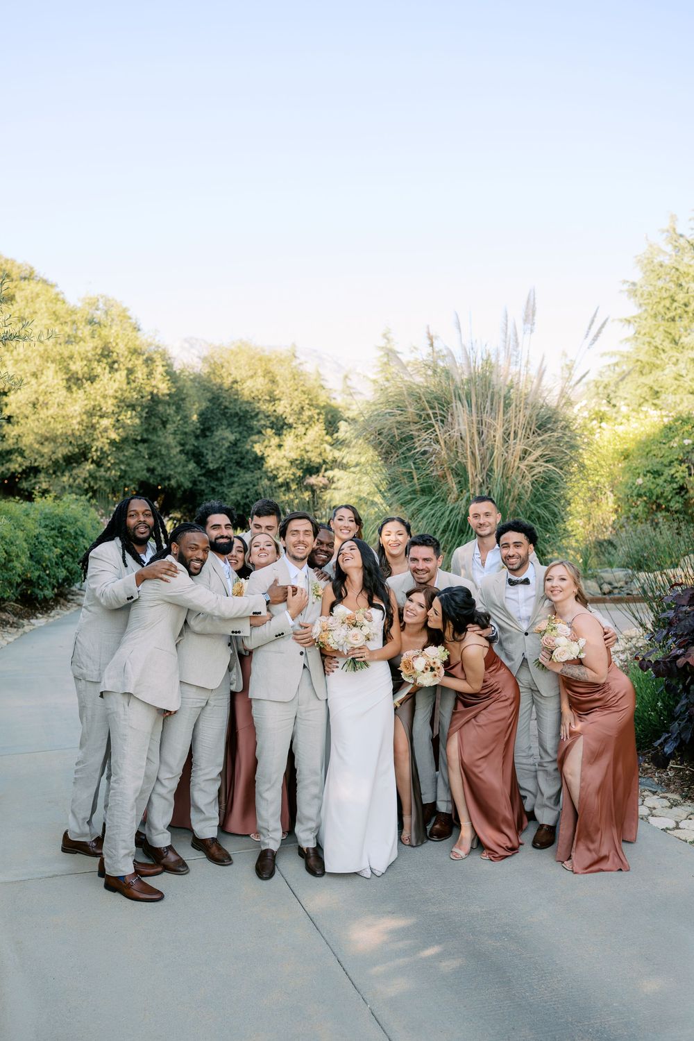 Wedding party poses outdoors in lush garden, groomsmen in gray suits and bridesmaids in terracotta dresses surrounding bride and groom.
