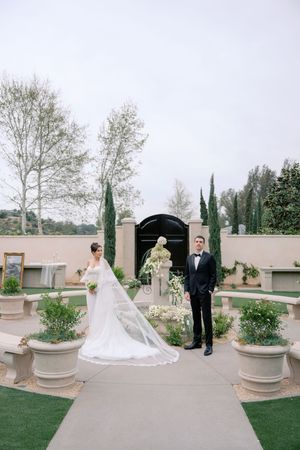 Bride in white gown with veil and groom in tuxedo stand in elegant outdoor garden wedding venue with greenery.