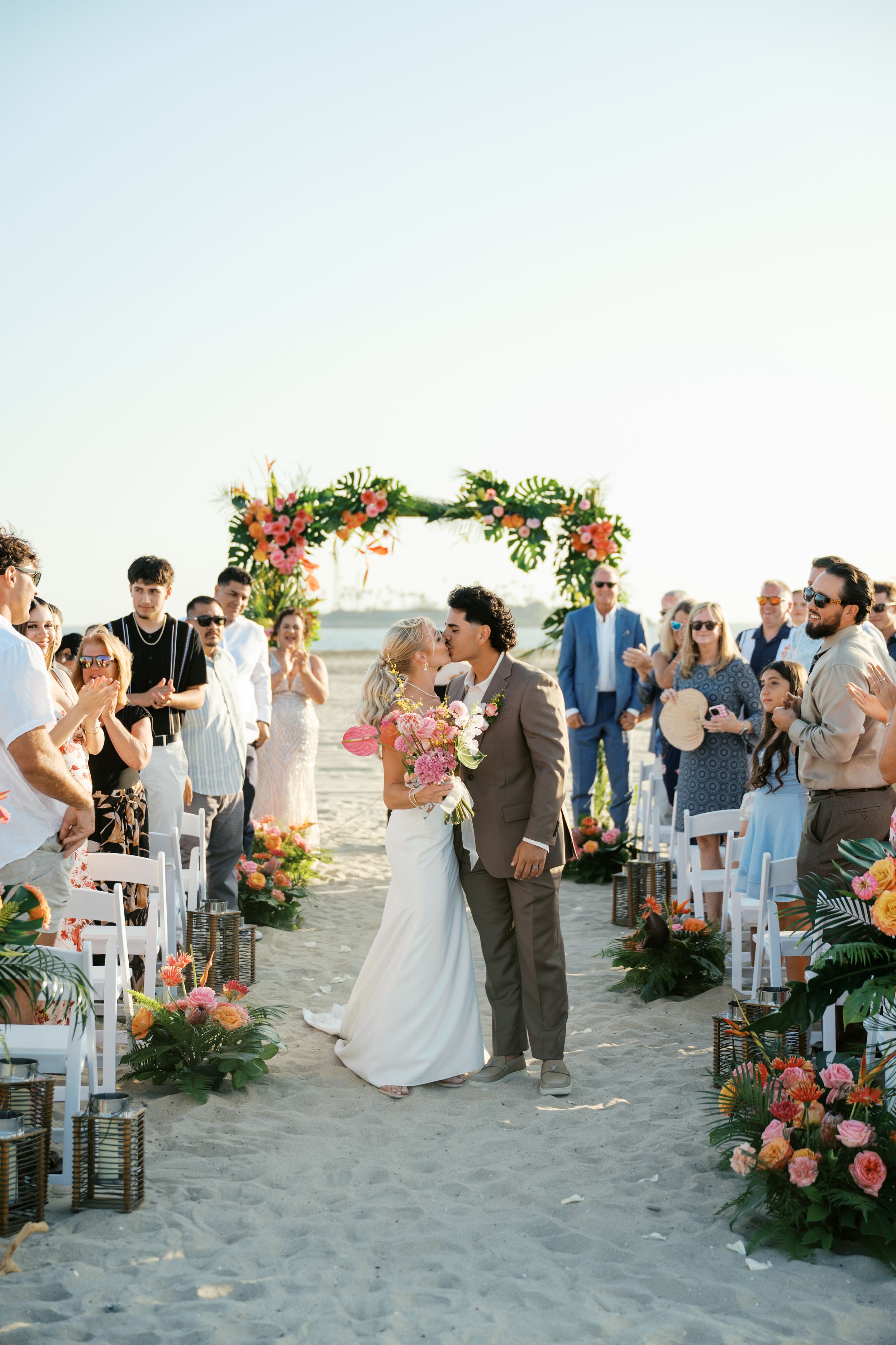 Beach wedding ceremony sequence showing guests gathered around floral arch and white chairs on sandy shoreline.