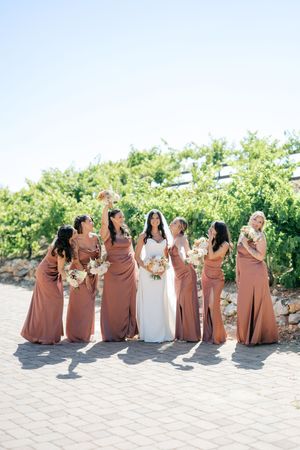 Bride in white gown poses with six bridesmaids in rust-colored satin dresses holding bouquets at a vineyard wedding.