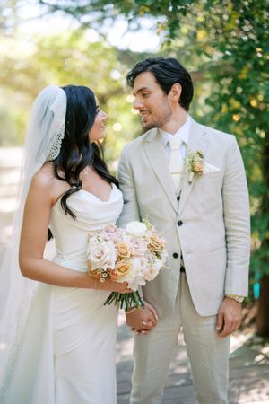Bride and groom gazing at each other outdoors, she holds a blush rose bouquet, he wears a beige suit with floral boutonniere.