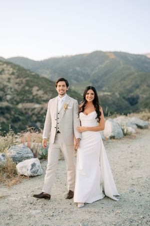 Bride and groom holding hands on a mountain trail, she in a white gown, he in a beige suit, with green hills behind.